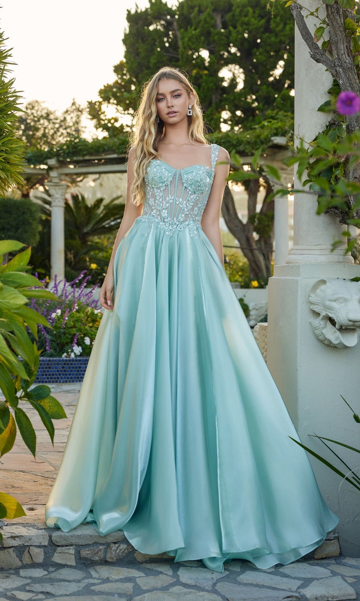 Outdoors in a garden, a woman wears the Juliet JT2639G long prom dress, featuring an elegant A-line silhouette with a corset bodice, delicate beaded applique, and a flowing light blue satin skirt.